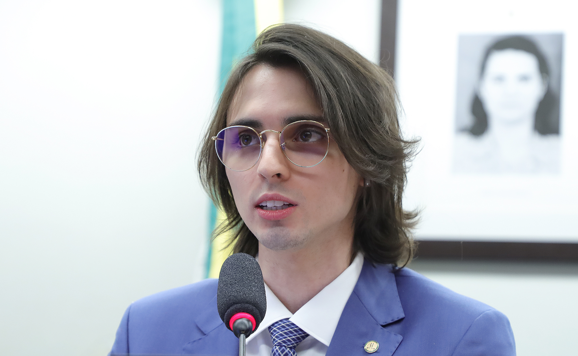 Amom de terno azul, falando ao microfone, com retrato ao fundo.